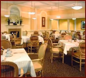 A dining room with multiple tables covered in white tablecloths, each set with cups and saucers. The room features cushioned chairs with wooden frames, a fireplace with a mantel decorated with plants and framed artwork, and soft lighting from ceiling fixtures.