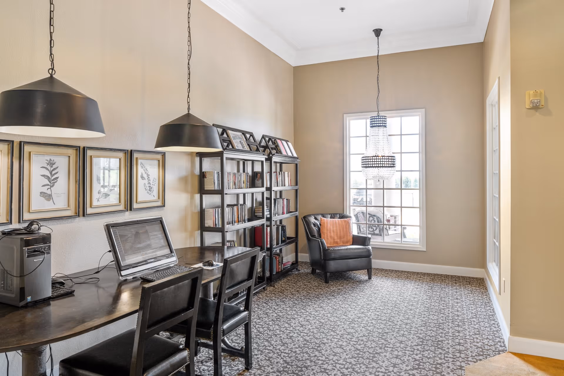 A cozy interior room with a long dark wooden table holding a desktop computer and two chairs. Above the table hang two large black pendant lights. On the wall behind the table are framed botanical prints. Next to the table is a black metal bookshelf filled with books and picture frames. A large window with multiple panes lets in natural light, illuminating a black leather armchair with an orange cushion beneath a hanging crystal chandelier. The room has beige walls and patterned carpet flooring.