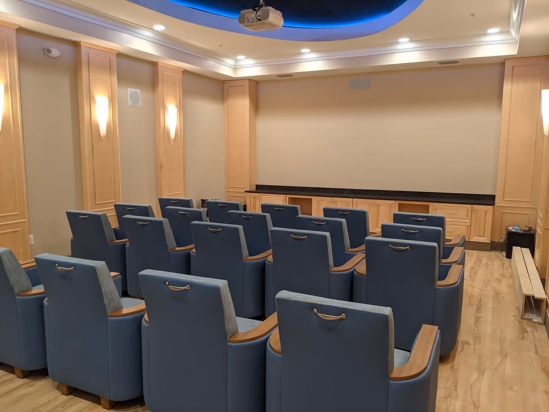 Small theater-style screening room with rows of blue upholstered seats facing a blank projection screen and ceiling projector.
