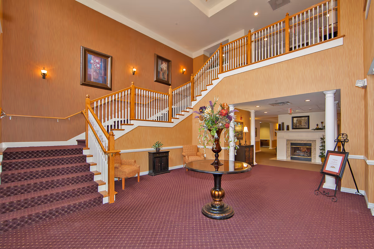Interior view of a senior living facility lobby with a carpeted floor and staircase with wooden handrails and white balusters. A round table with a large floral arrangement is centered in the room. Two wicker chairs and a small cabinet are placed against the wall. The walls are decorated with framed floral paintings and wall sconces. In the background, there is a fireplace with a mantel and a framed picture above it, flanked by two white columns.