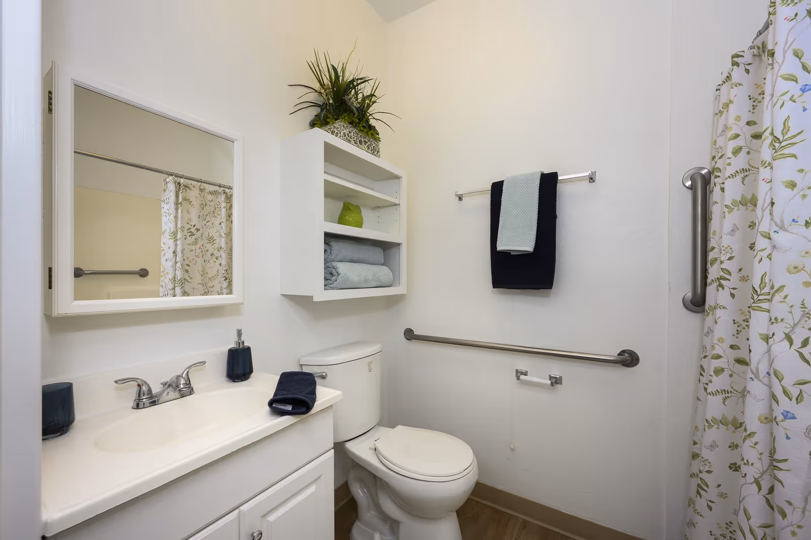 A clean and well-lit bathroom with a white vanity and sink, a mirror above the sink, a toilet with a white seat, and a white wall-mounted cabinet holding folded towels and a green decorative item. There are two towel bars, one with two folded towels and the other empty, and a floral-patterned shower curtain partially visible on the right side. The bathroom has grab bars for accessibility and a wooden floor.