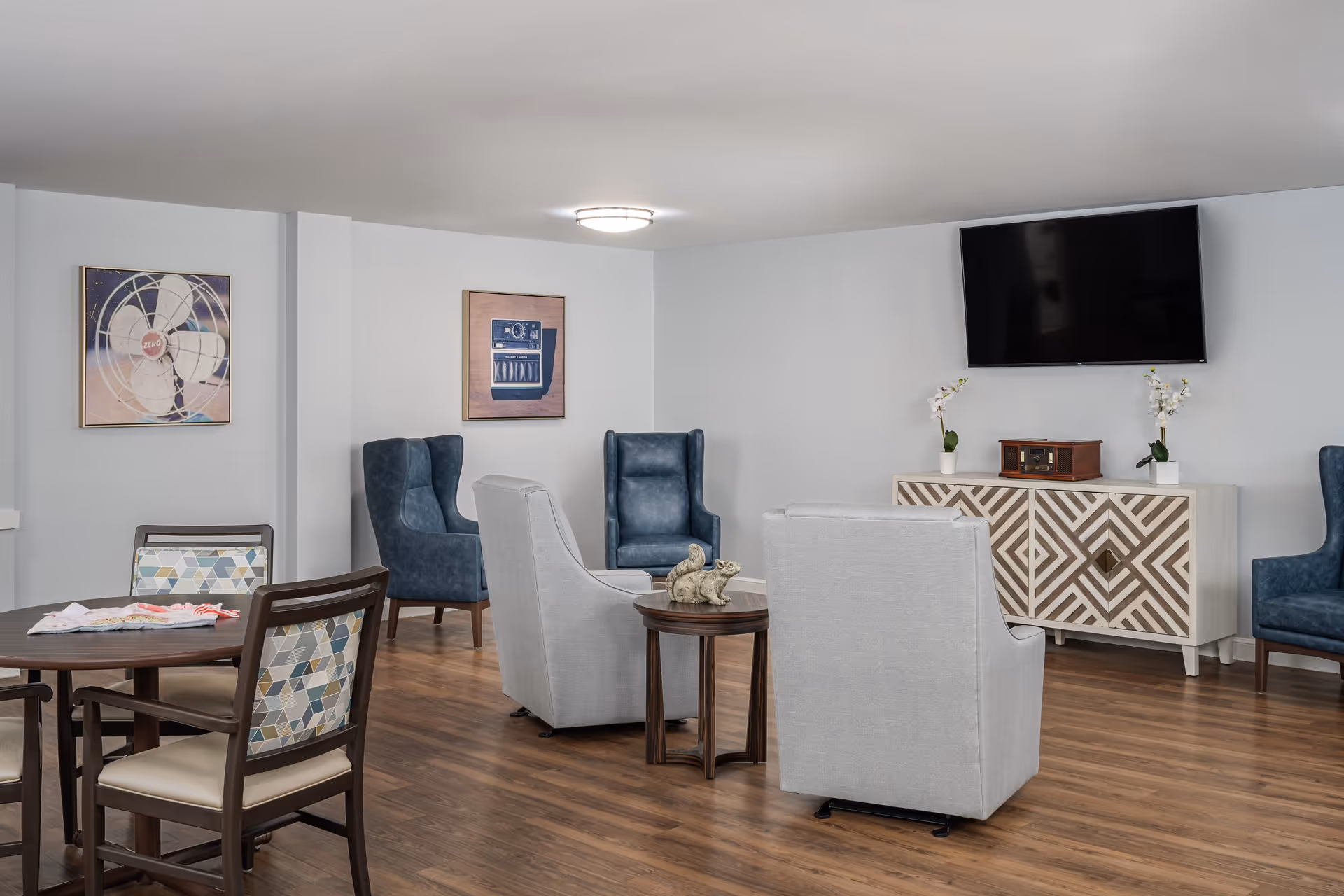 A bright common room with upholstered armchairs, a table and chairs, decorative cabinet, wall art, and a wall-mounted TV.