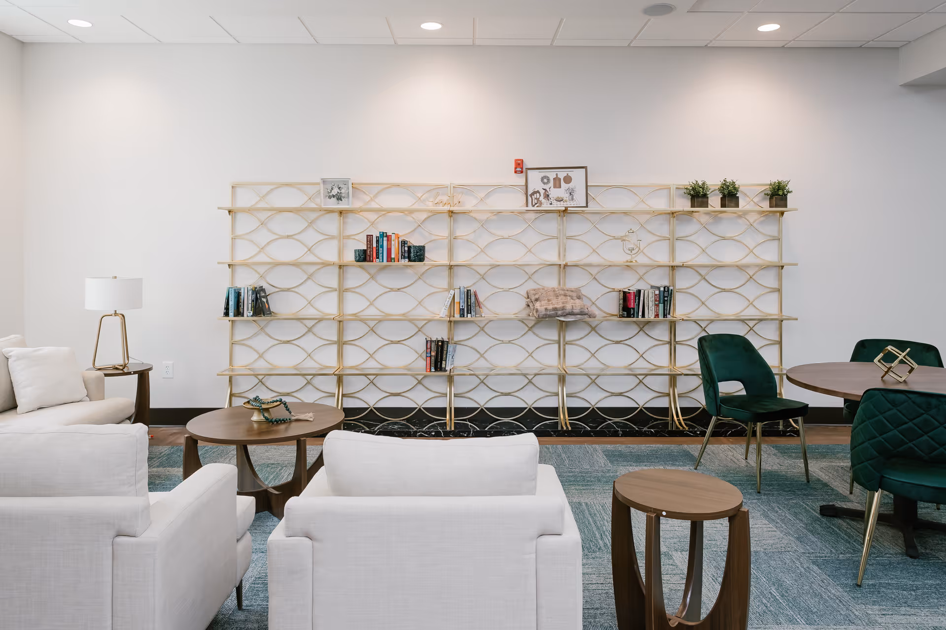 A modern senior living common area with white cushioned armchairs, a wooden coffee table, a side table with a lamp, and a decorative gold metal bookshelf against a white wall. The bookshelf holds a few books, small plants, and framed pictures. There are also green upholstered chairs around a round wooden table on a blue patterned carpet.