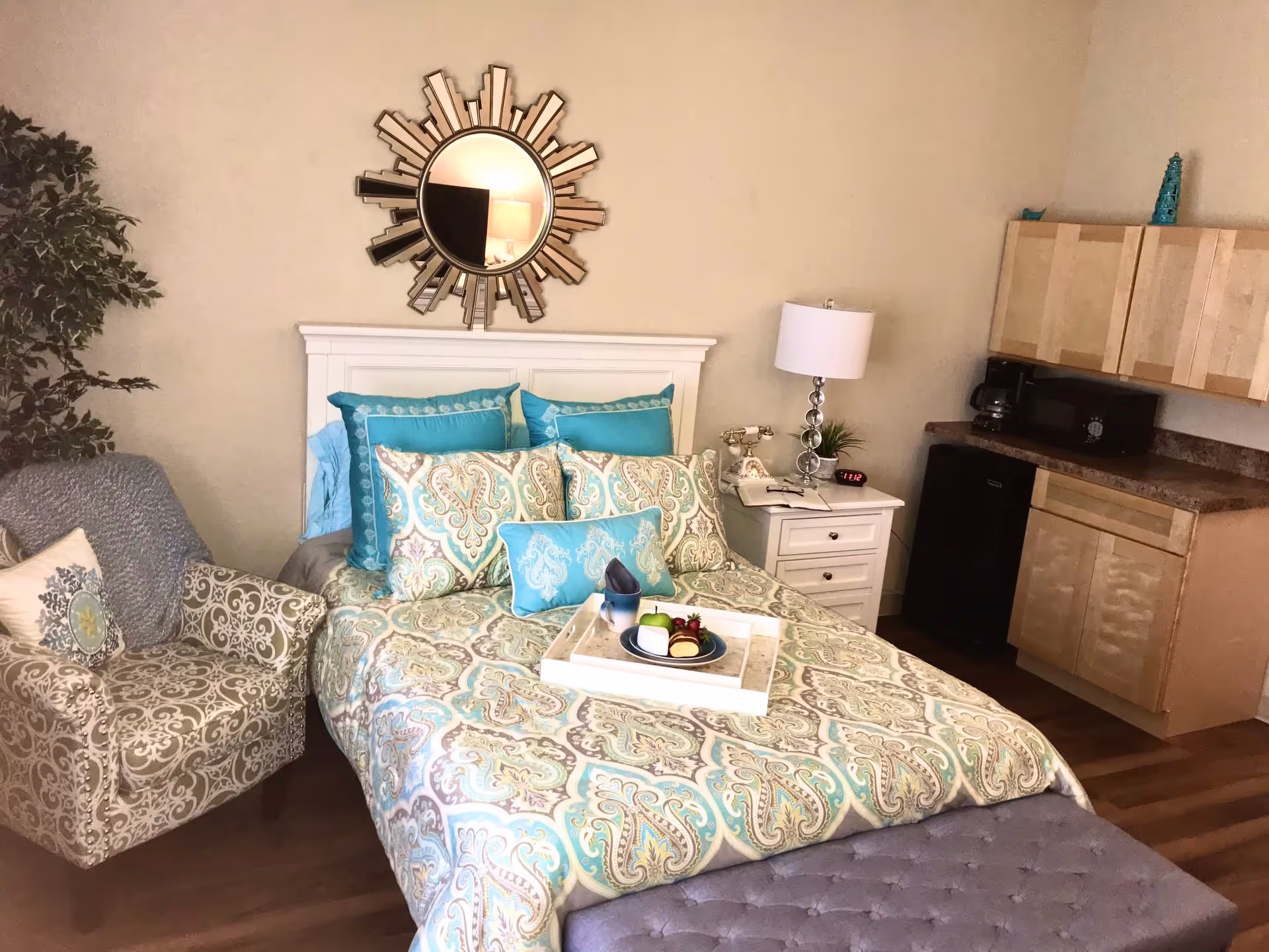 A cozy bedroom with a bed featuring a patterned bedspread and multiple pillows in shades of turquoise and beige. A white headboard is mounted on the wall behind the bed, with a decorative sunburst mirror above it. To the right of the bed is a white nightstand with a lamp, an old-fashioned telephone, a small plant, and a digital clock. Next to the nightstand is a kitchenette area with light wood cabinets, a microwave, a coffee maker, and a mini refrigerator. To the left of the bed is a patterned armchair with a cushion and a throw blanket. The floor is wooden, and the overall room has a warm and inviting atmosphere.