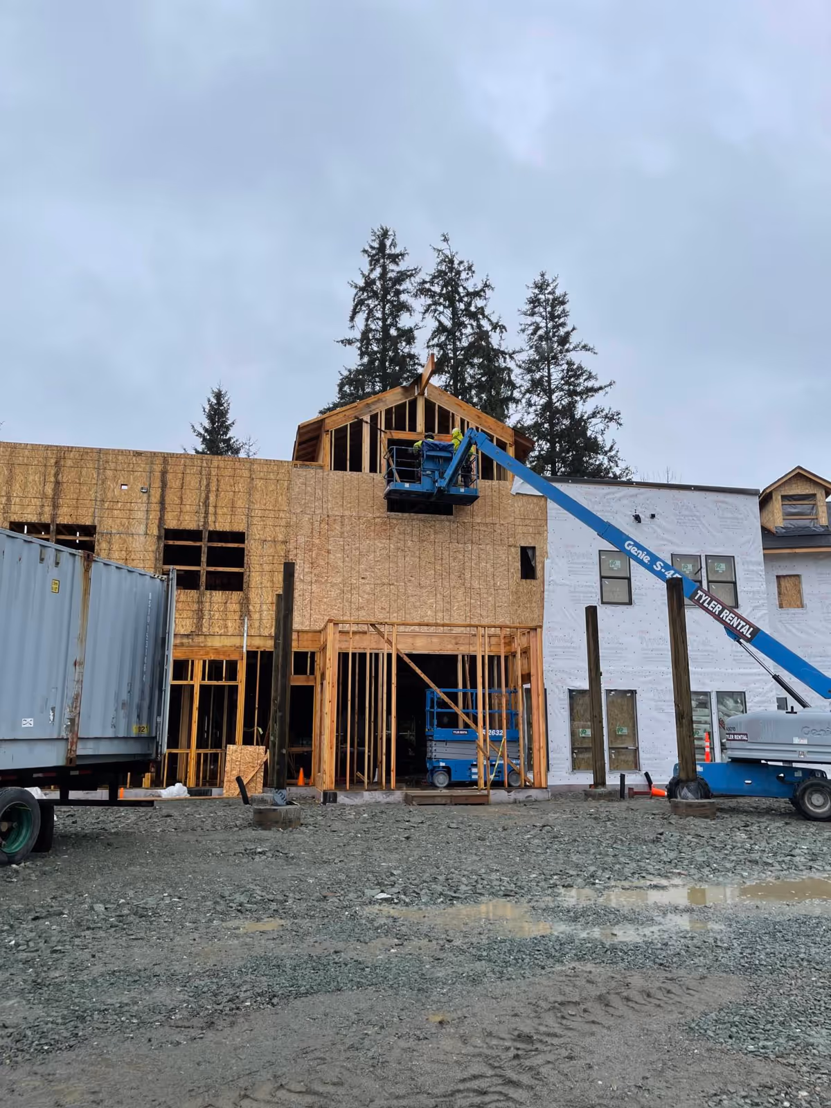 Front view of a multi-story building under construction with exposed wood framing, a blue boom lift, and a gravel lot in front.
