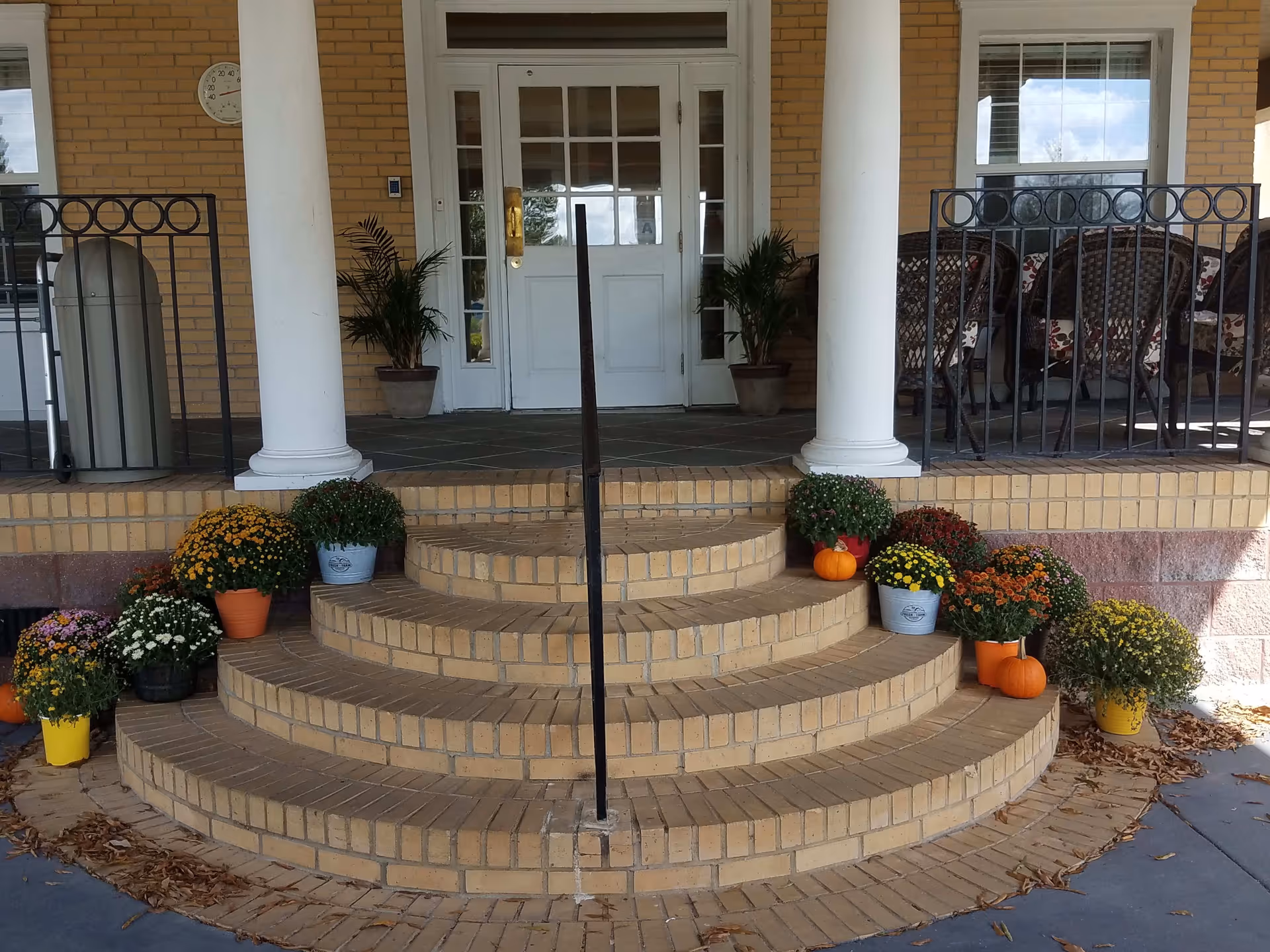 Brick semicircular front steps decorated with potted mums and small pumpkins leading up to a white double door flanked by columns.