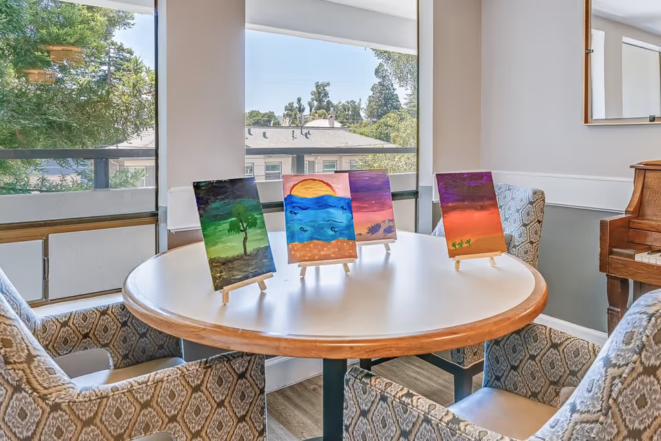 A round wooden table with four small paintings on easels displayed on top. The table is surrounded by four patterned armchairs. Large windows in the background show trees and a building outside. A piano is partially visible on the right side of the image.