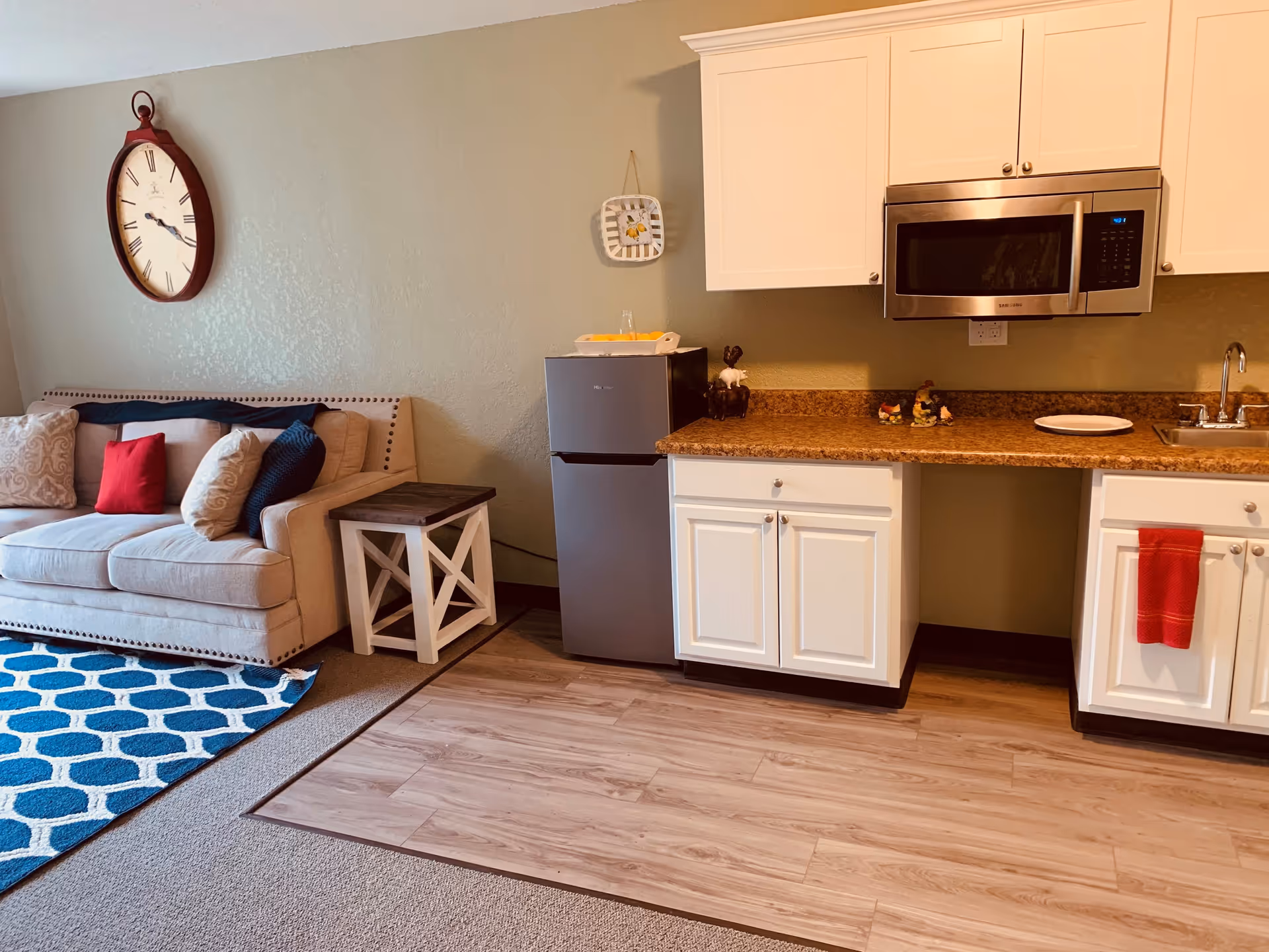 Interior view of a small living area and kitchenette in a retirement community. The living area has a beige couch with multiple cushions in red, beige, and dark blue, a small wooden side table, and a blue patterned rug on the carpeted floor. The kitchenette features white cabinets, a small stainless steel refrigerator, a microwave, a sink, and a countertop with decorative items and a red towel hanging from one cabinet. A large round wall clock is mounted on the wall above the couch.