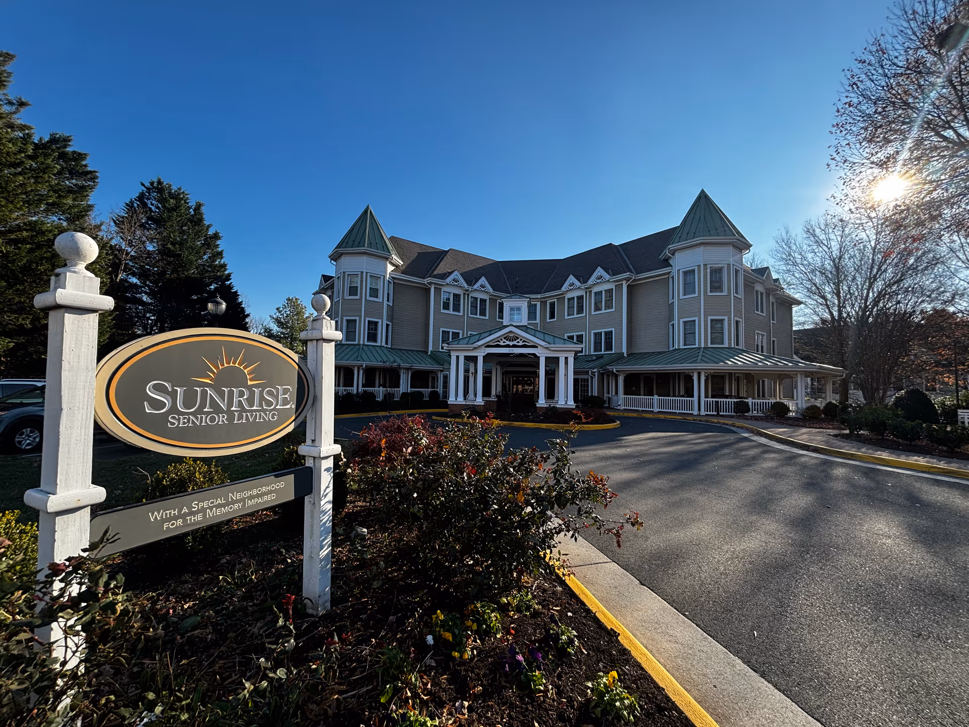 A three-story Sunrise Senior Living building with a covered entrance, circular driveway, and a sign in the foreground under a clear blue sky.