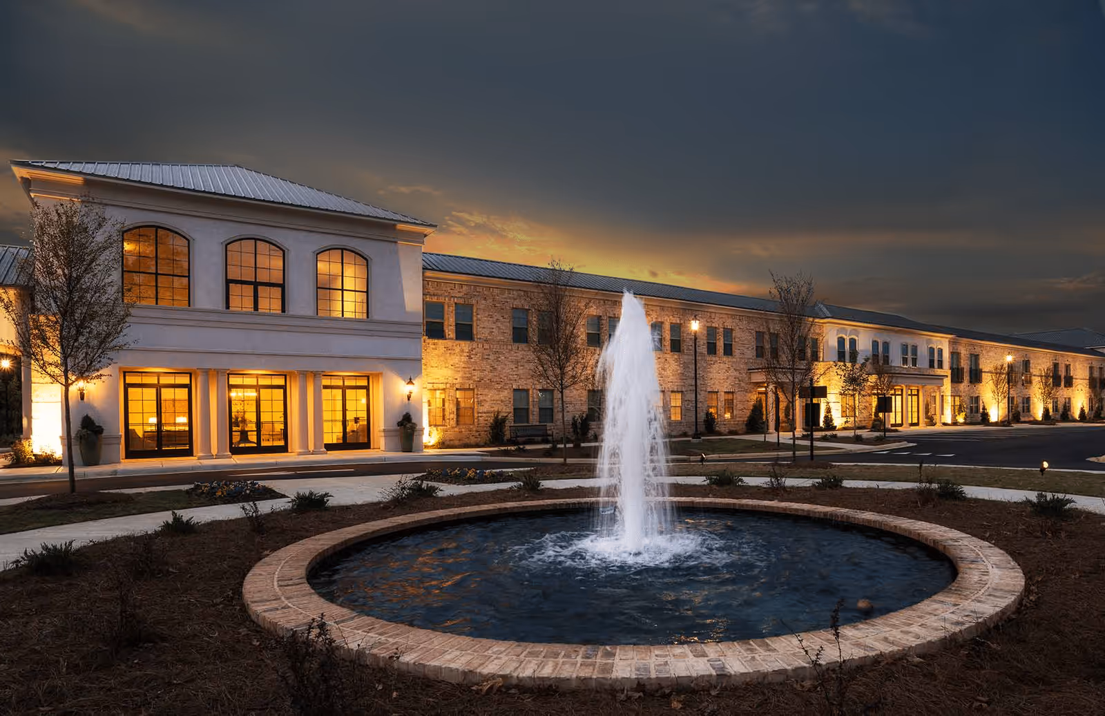 Exterior view of The Blake at Flowood senior living facility at dusk, featuring a large two-story building with illuminated windows and a central water fountain in a circular brick basin in the foreground.