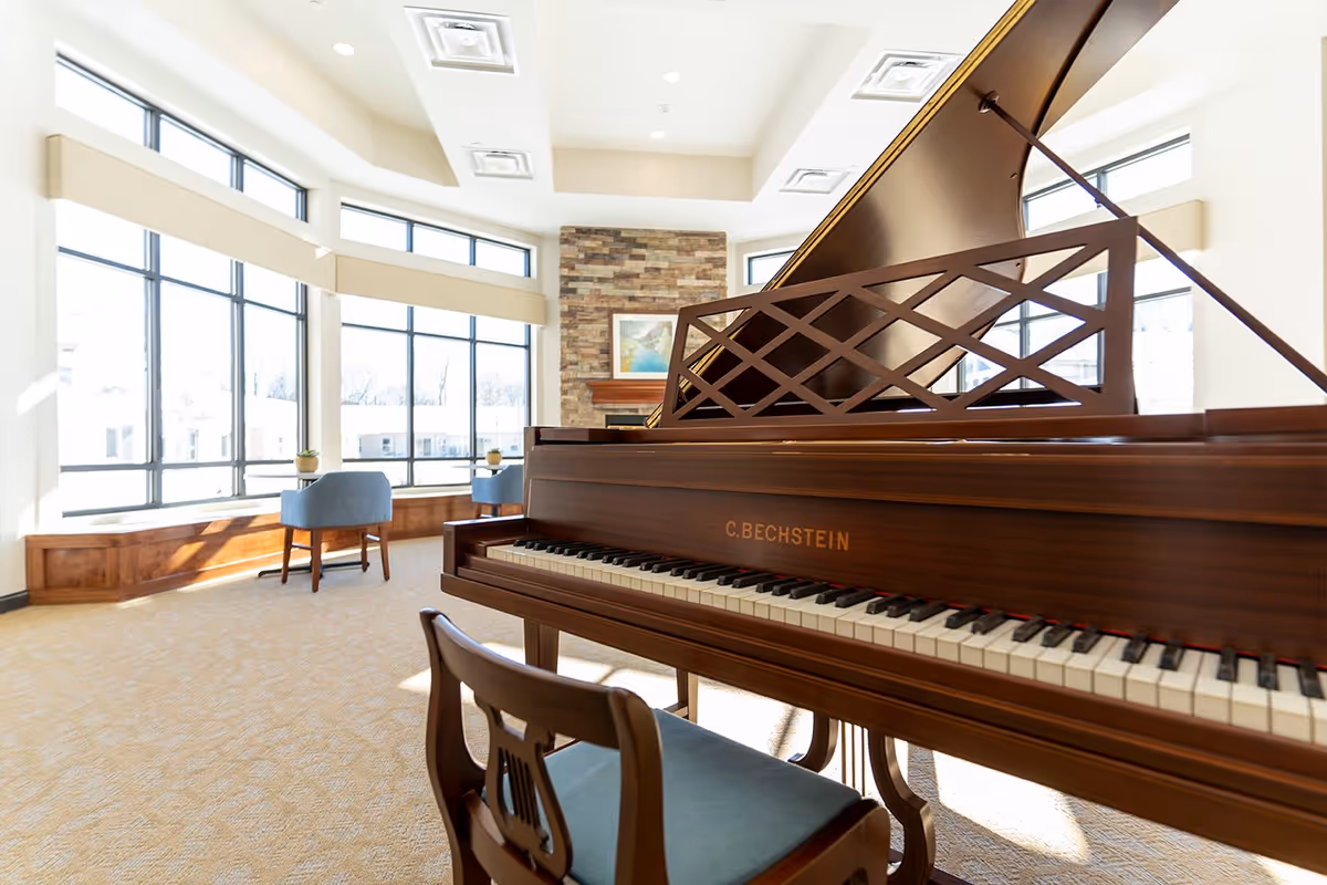Bright room with large windows, a grand piano with a wooden chair, two blue armchairs near the windows, and a stone fireplace with a framed picture above it.