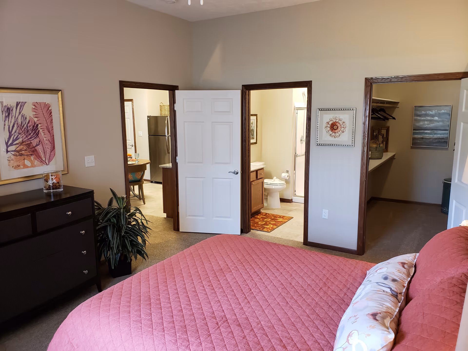 Cozy bedroom with a pink quilted bed, dresser and plant, with open doors revealing a bathroom, kitchen and walk-in closet.