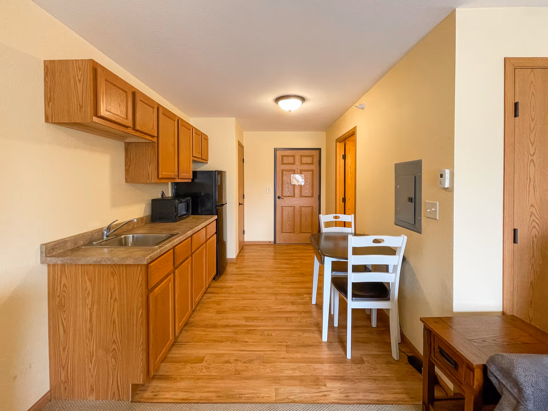 Interior view of a small kitchen and dining area in a senior living facility. The kitchen has wooden cabinets, a countertop with a sink, a microwave, and a black refrigerator. The dining area features a small dark wooden table with two white chairs. The floor is wood laminate, and there are wooden doors and light beige walls.
