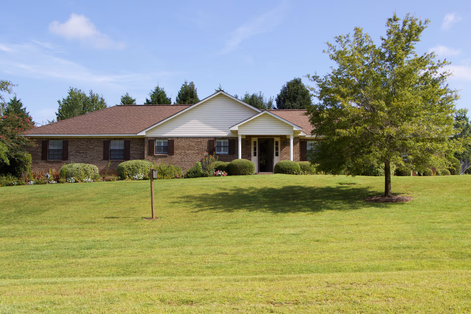 Single-story brick building with a brown roof and white trim, surrounded by green grass and bushes, with a tree on the right side under a blue sky with some clouds.