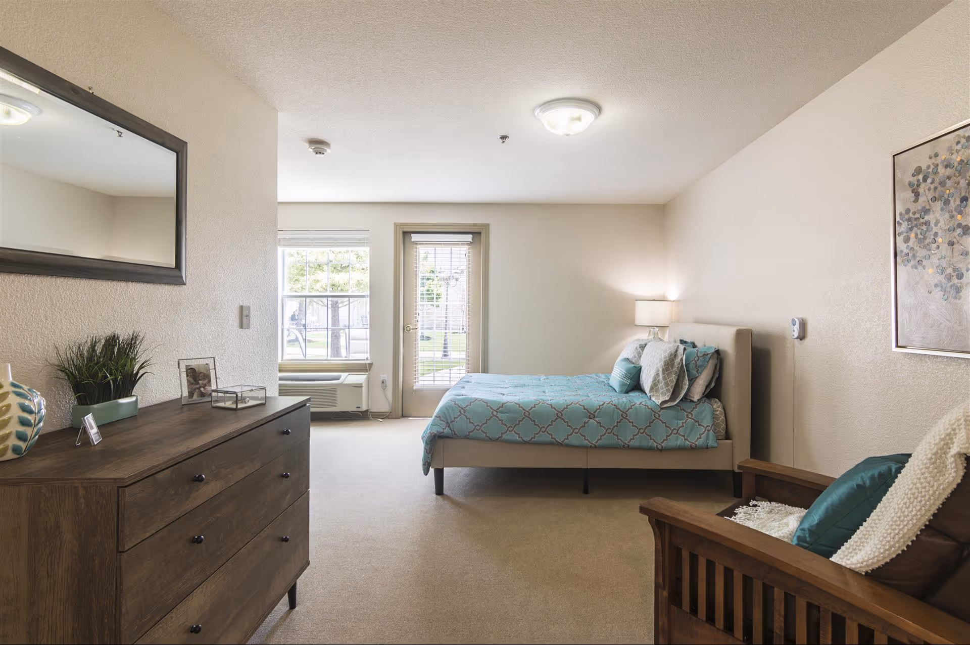 A bright, neatly arranged bedroom in an assisted living facility featuring a bed with blue patterned bedding, a wooden dresser with decorative items, a wooden armchair with cushions and a throw, a window and a glass door letting in natural light, and a framed artwork on the wall.
