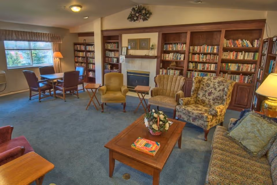 A cozy living room area in a senior living facility featuring a variety of upholstered chairs and a sofa arranged around a wooden coffee table with a floral centerpiece and books. Behind the seating area is a fireplace flanked by built-in wooden bookshelves filled with books. A window with curtains and a table with four chairs are visible in the background, along with a standing lamp and a table lamp providing warm lighting.