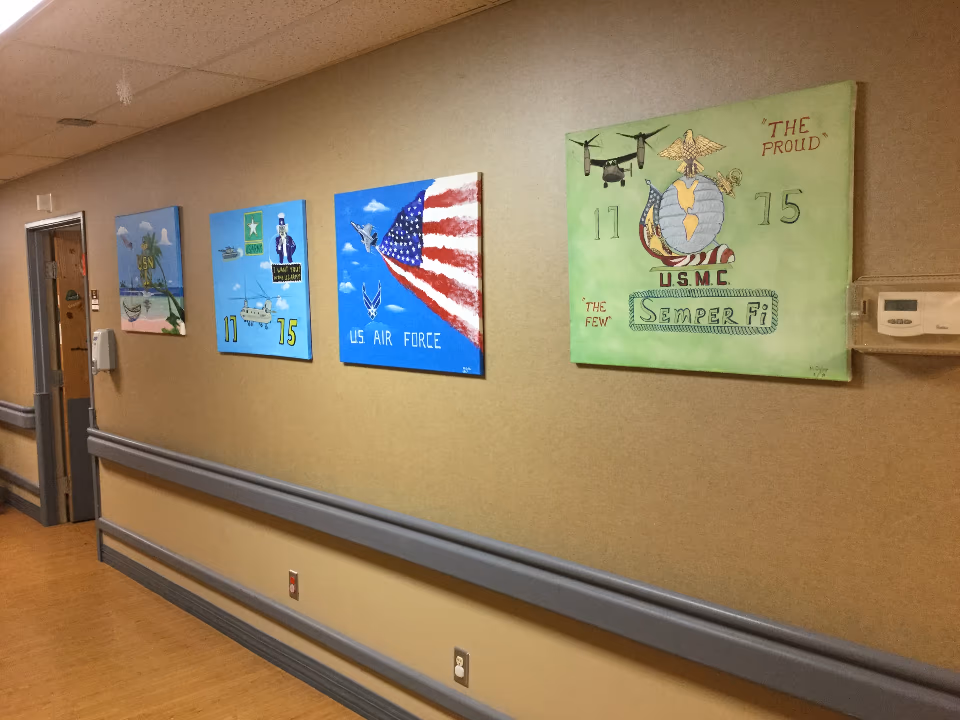 A hallway in a senior living facility with beige walls and wooden flooring. Four paintings are displayed on the wall, each representing different branches of the U.S. military including the Navy, Army, Air Force, and Marine Corps. The hallway has handrails along the walls and a thermostat on the right side.