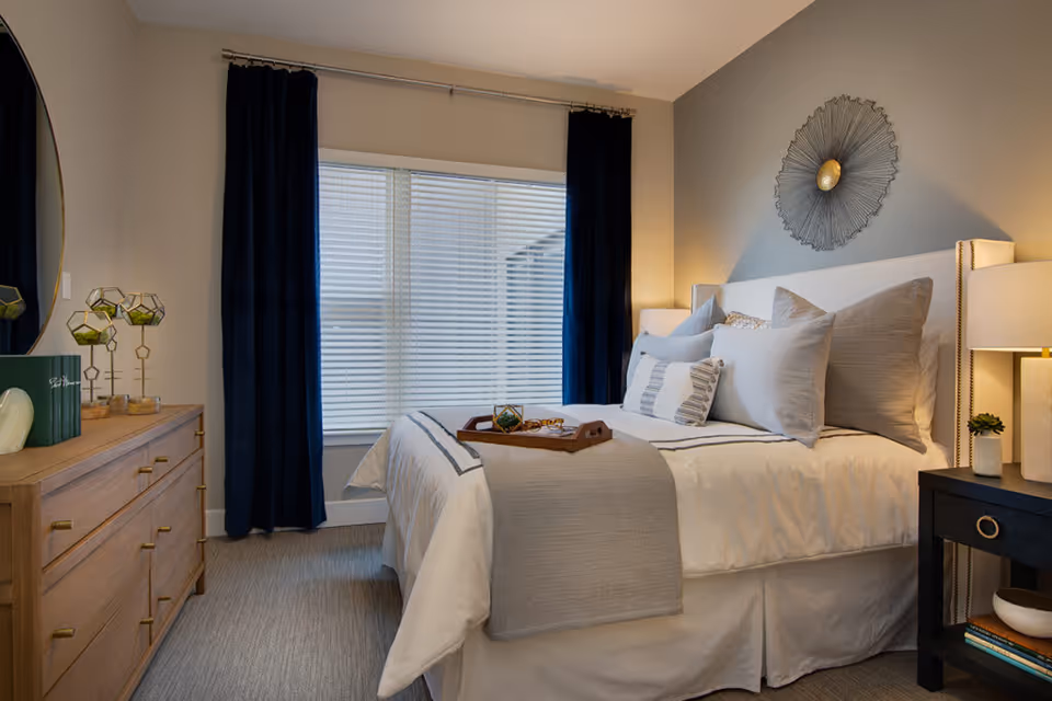 A neatly made bed with multiple pillows and a gray throw blanket in a bedroom. There is a wooden dresser with decorative items on the left and a black nightstand with a lamp and small plant on the right. A large window with white blinds and dark blue curtains is behind the bed, and a decorative wall piece hangs above the headboard.