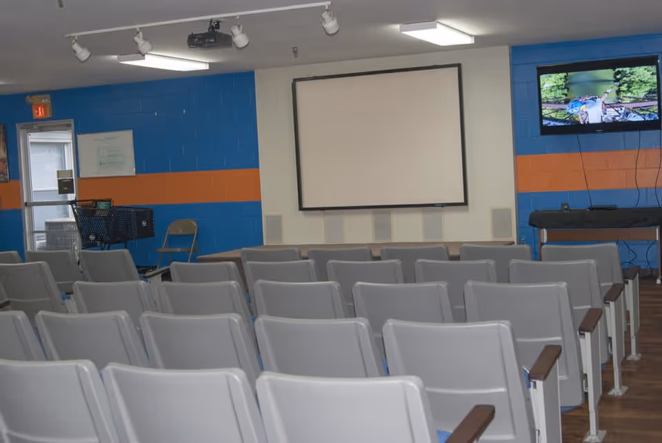 A room with multiple rows of gray chairs facing a large white projection screen mounted on a wall painted blue with an orange stripe. There is a television screen on the right side showing an outdoor scene, a folding chair, a shopping cart, and a door with an exit sign above it on the left side.