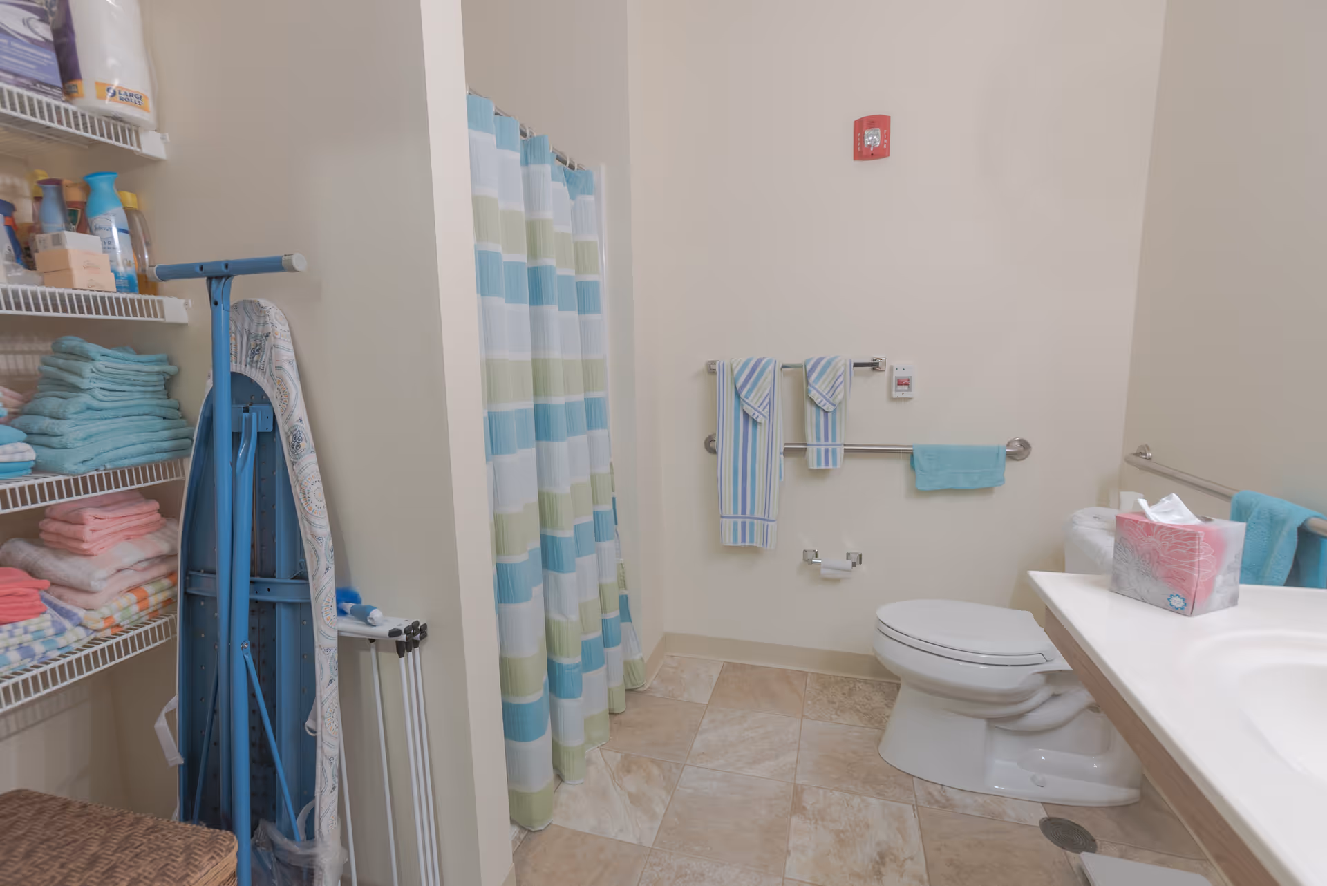 A clean bathroom with a toilet, a sink with a tissue box on the counter, and a shower with a striped blue, green, and white curtain. There are several towels hanging on wall-mounted bars and shelves stocked with folded towels and various toiletries. An ironing board and an iron are also visible in the corner.