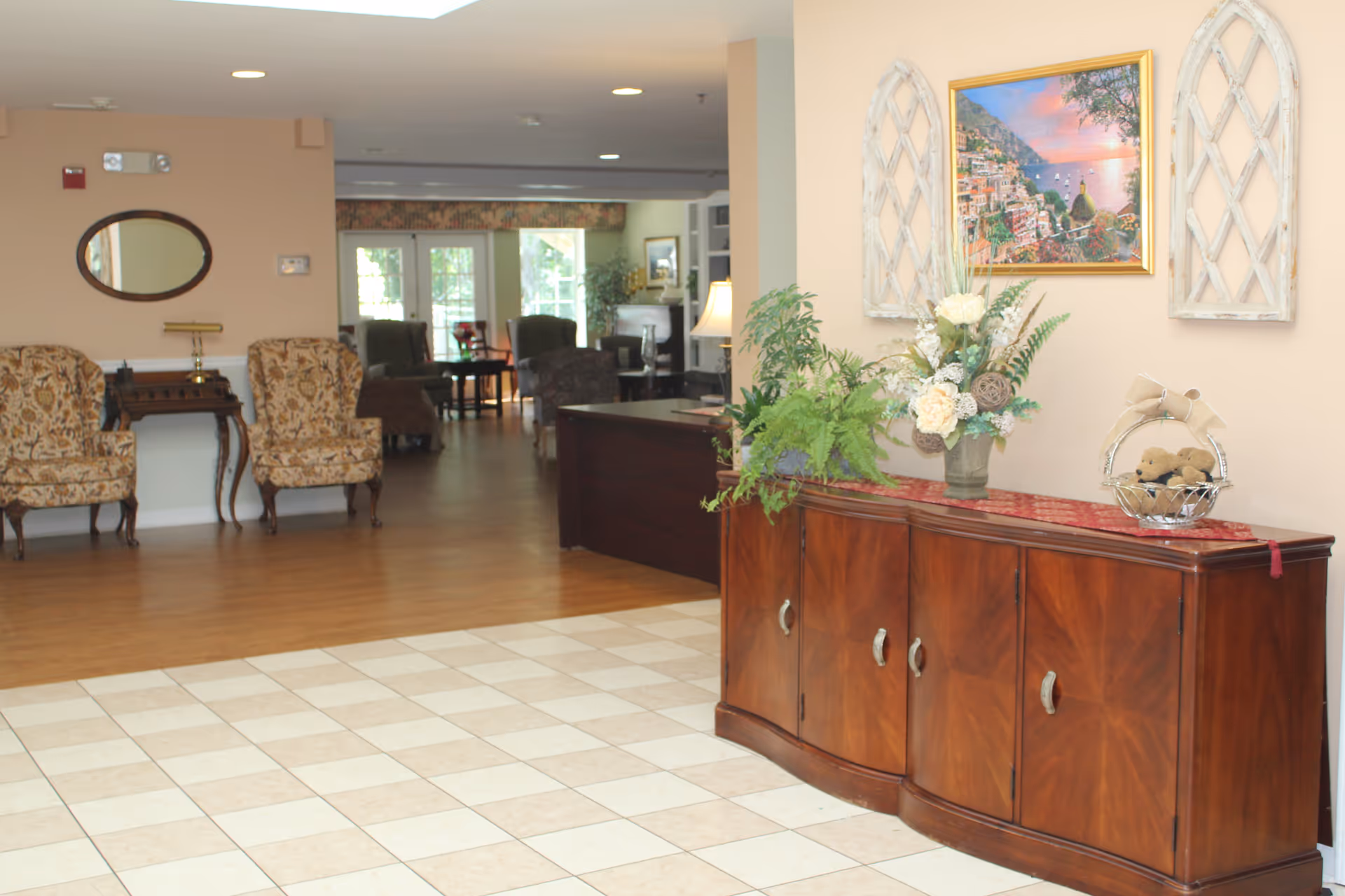 Interior view of a senior living facility featuring a wooden sideboard with decorative plants and a basket on top, a painting and two decorative wall pieces above it. In the background, there are patterned armchairs, a small table with a lamp, and a seating area with more chairs and tables near large windows letting in natural light.