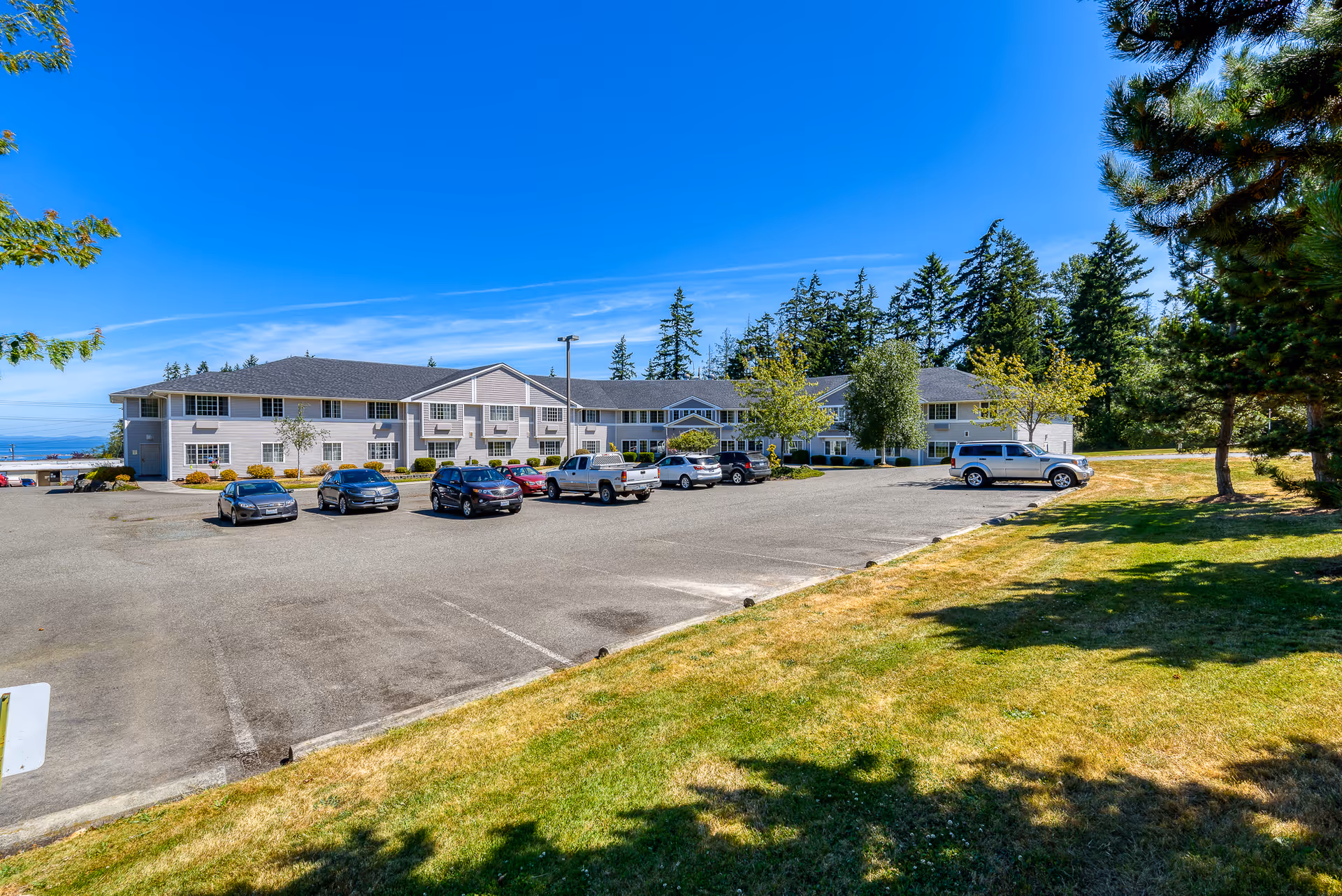 Exterior view of a two-story assisted living facility building with a parking lot in front containing several parked cars. The building is surrounded by trees and greenery under a clear blue sky.
