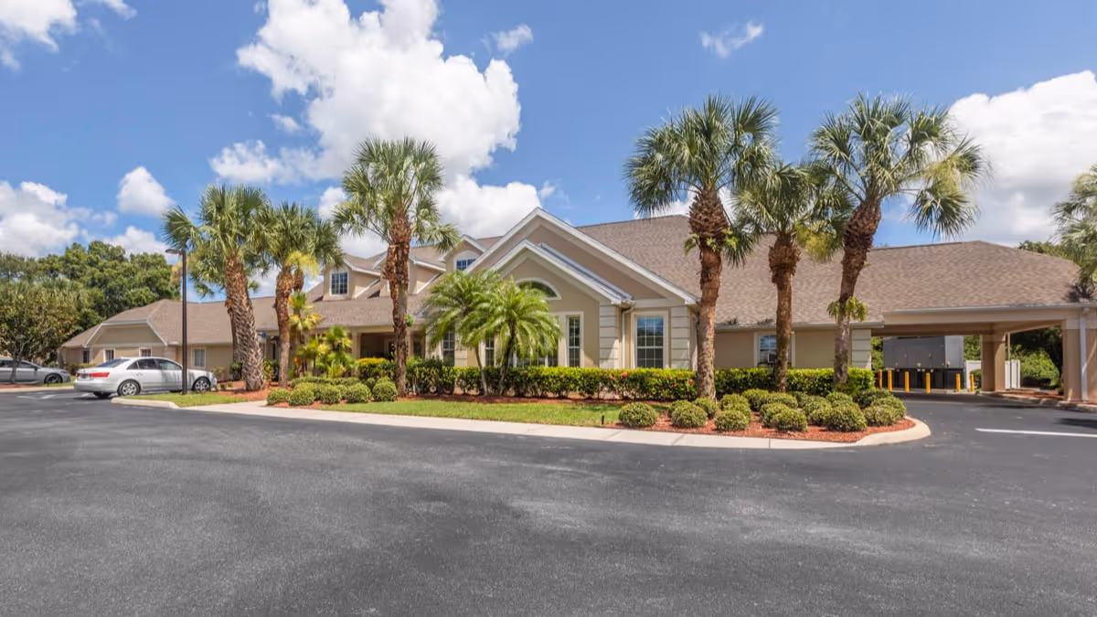 Exterior view of a single-story building with a sloped roof, surrounded by palm trees and well-maintained landscaping under a partly cloudy blue sky. There is a paved driveway and parking area in front of the building.
