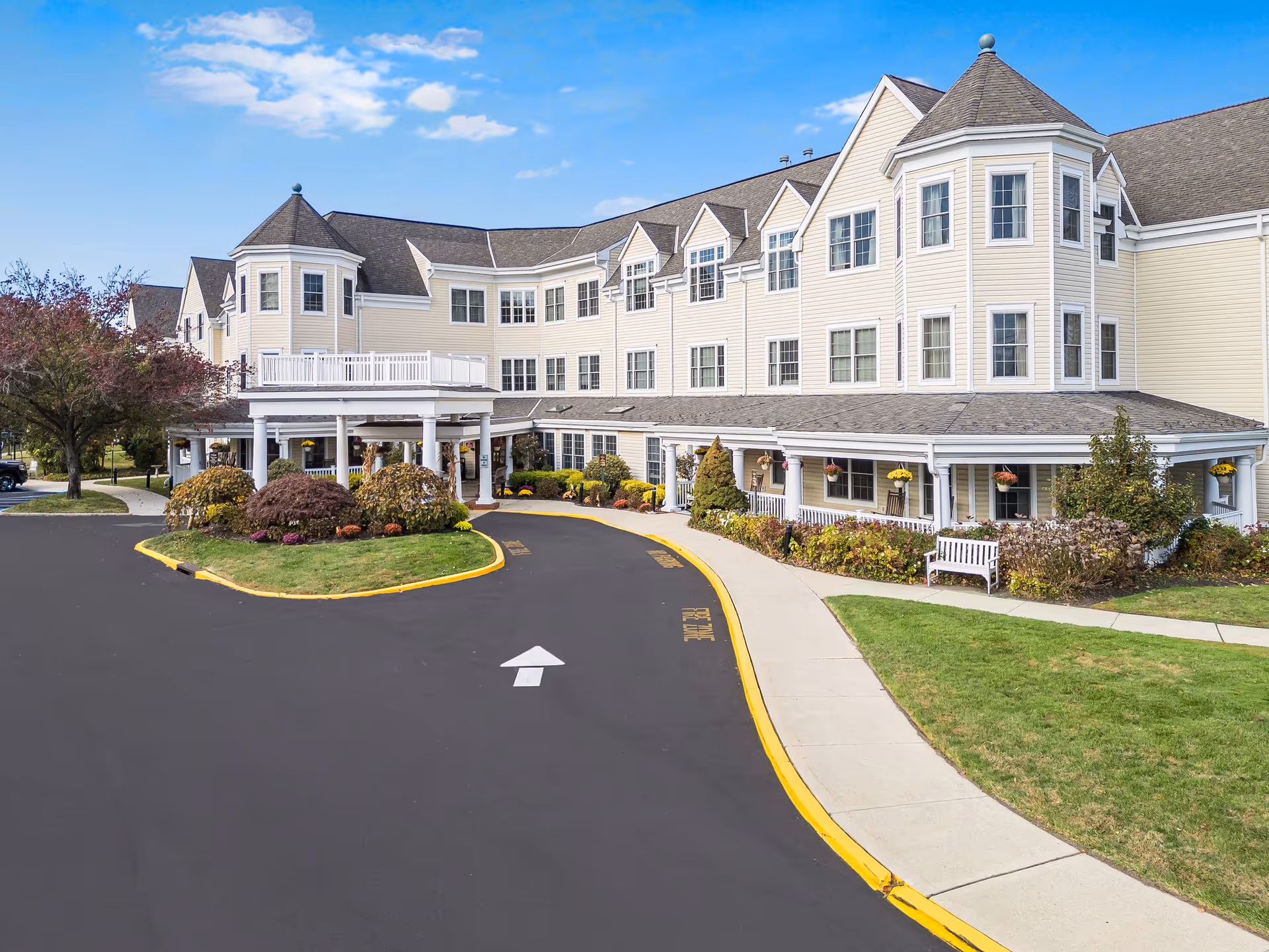 Front exterior of a pale-yellow multi-story senior living building with a covered entrance, circular driveway, and landscaped grounds.