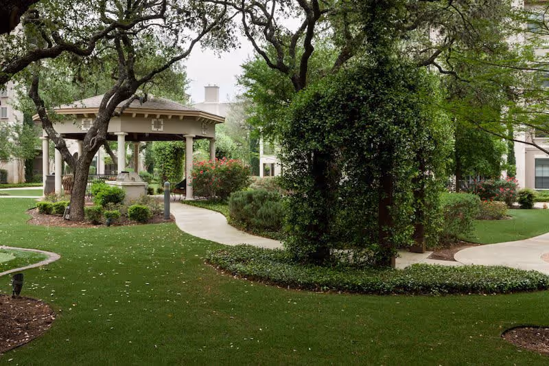 A well-maintained outdoor garden area with green grass, trees, shrubs, and a covered gazebo structure with columns. Curved concrete pathways wind through the garden, surrounded by various plants and bushes.