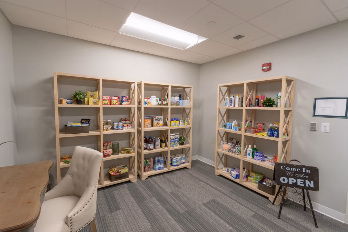Small interior room with wooden shelving stocked with snacks, beverages, and toiletries, plus a chair and desk.