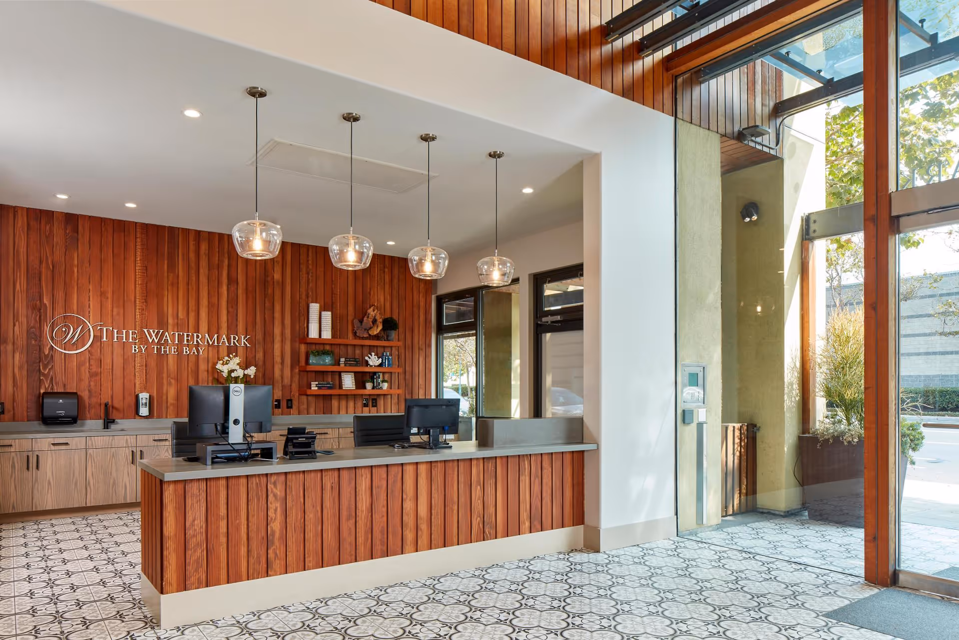 Reception area with a wooden front desk and wood-paneled wall behind it. The desk has two computer monitors and office equipment. Three pendant lights hang from the ceiling. There are shelves with decorative items and books on the wall. Large glass windows and doors allow natural light to enter the space.