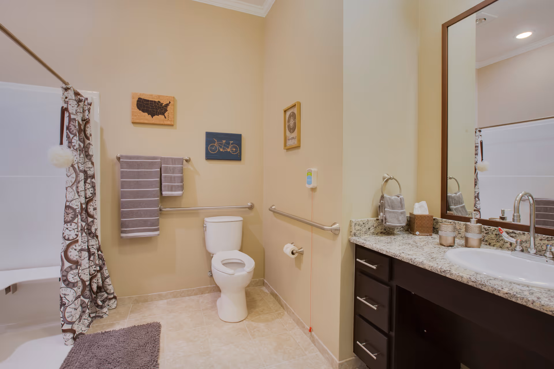 A clean and accessible bathroom with beige walls and tiled floor. The bathroom features a white toilet with grab bars on both sides, a shower with a curtain decorated with clock faces, a gray bath mat, and a granite countertop with a sink, faucet, and various toiletries. There are three framed pictures on the walls and gray towels hanging on a towel rack.