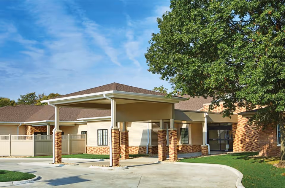 Front entrance of a single-story senior living building with a covered drive-up canopy, brick accents and surrounding trees.