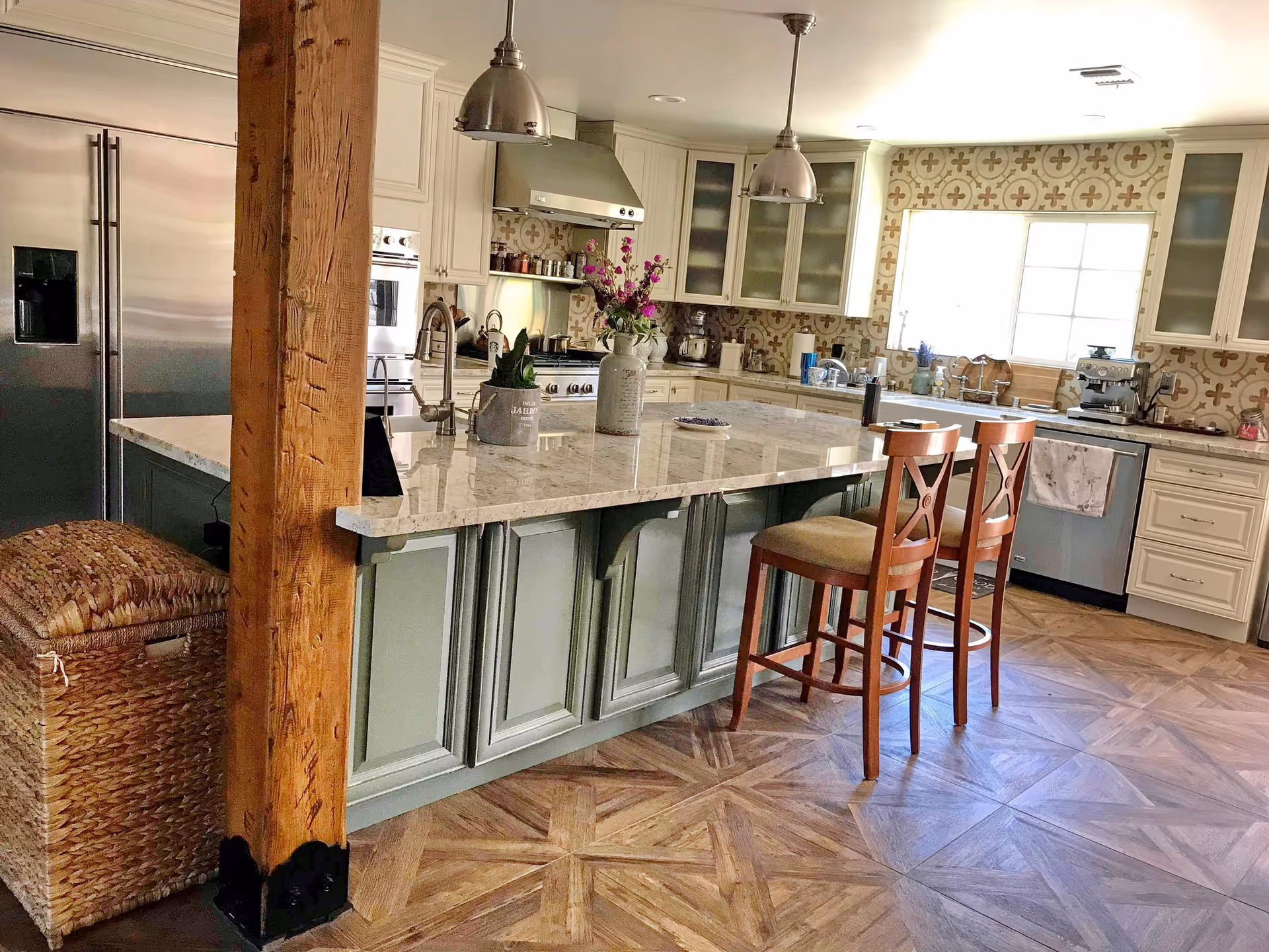 A spacious kitchen with a large island countertop featuring a marble surface and two wooden chairs. The kitchen has cream-colored cabinets, a stainless steel refrigerator, oven, and dishwasher. There are pendant lights hanging from the ceiling, a window above the sink, and decorative patterned tiles on the backsplash. A wooden support beam and a woven basket are visible on the left side.