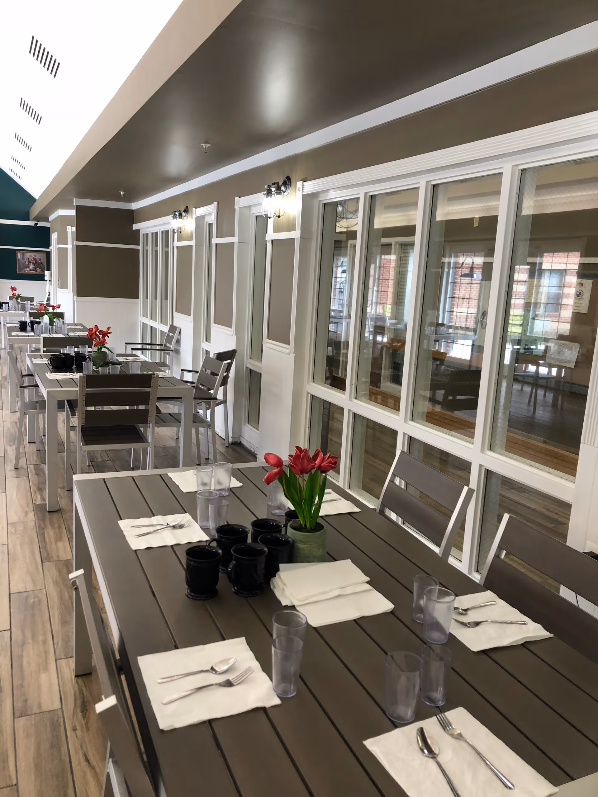 A dining area with multiple tables and chairs arranged in a row along a wall with large windows. Each table is set with napkins, utensils, plastic cups, and black mugs. A small vase with red flowers is placed on the nearest table. The room has wood-patterned flooring and neutral-colored walls with white trim.