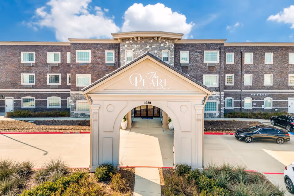 Front exterior view of The Pearl Senior Living at Lewisville building with a covered entrance structure, multiple windows, parked cars, and landscaped bushes under a blue sky with some clouds.