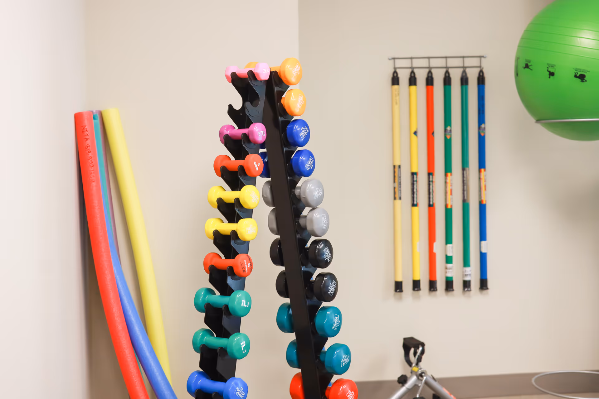 A fitness room with a rack of colorful dumbbells, several foam pool noodles leaning against the wall, a set of colorful exercise sticks hanging on a wall-mounted rack, and a large green exercise ball partially visible on the right.
