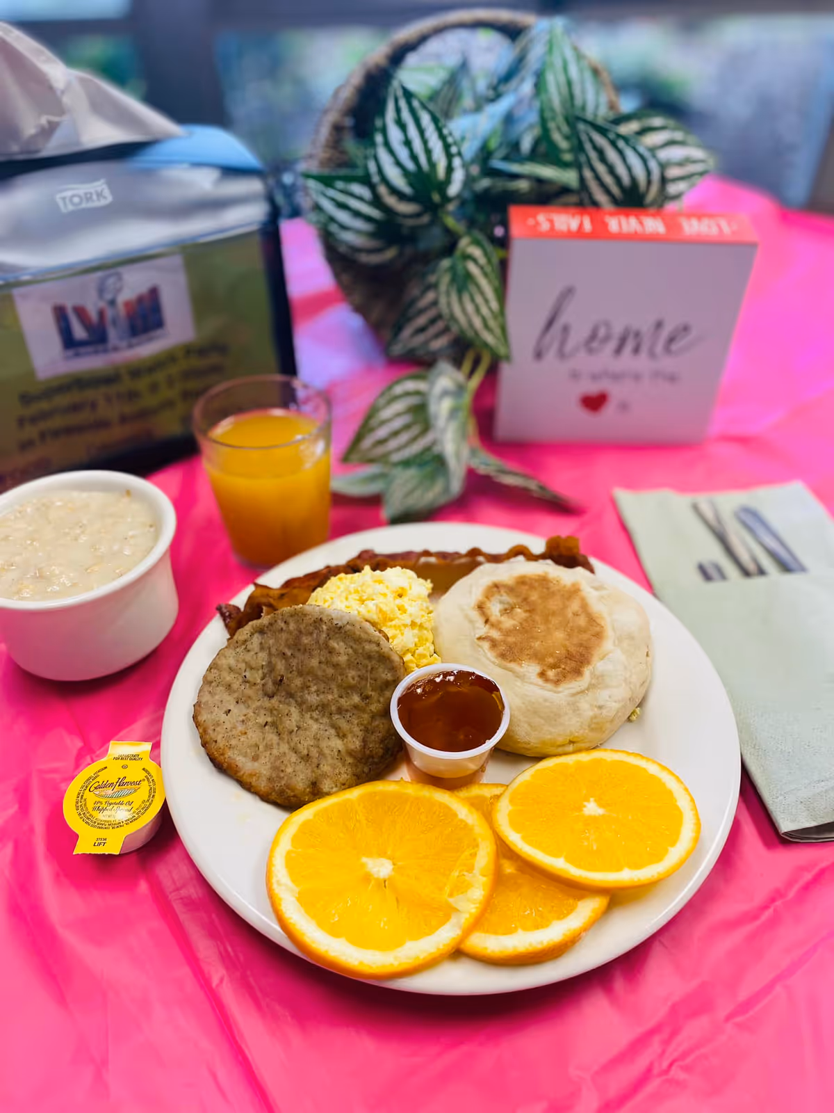 A breakfast plate with scrambled eggs, sausage patty, an English muffin, three orange slices, and a small cup of jelly. Next to the plate is a cup of oatmeal, a small container of butter, a glass of orange juice, and a napkin with utensils. In the background, there is a plant and a decorative block with the word 'home' on it, all set on a pink tablecloth.