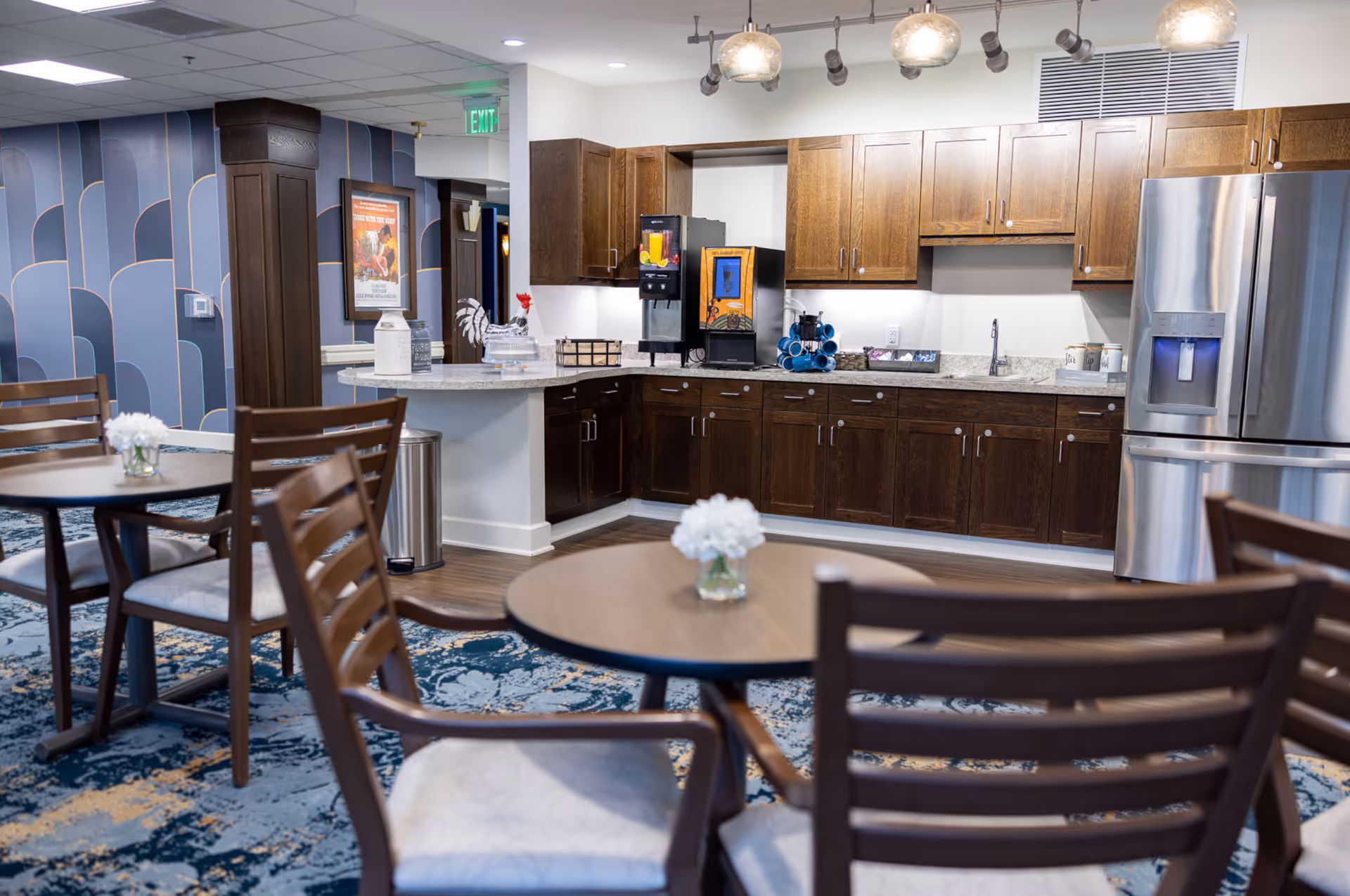A cozy kitchen and dining area in a senior living facility featuring wooden cabinets, a stainless steel refrigerator, a beverage dispenser, and round tables with wooden chairs. The room has a patterned blue carpet and decorative lighting fixtures.