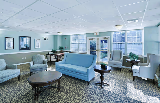 A bright and spacious living room area with light blue walls and carpeted floor featuring a patterned design. The room contains a light blue sofa, several armchairs, wooden coffee tables, and side tables with plants. Large windows and glass doors allow natural light to fill the space.