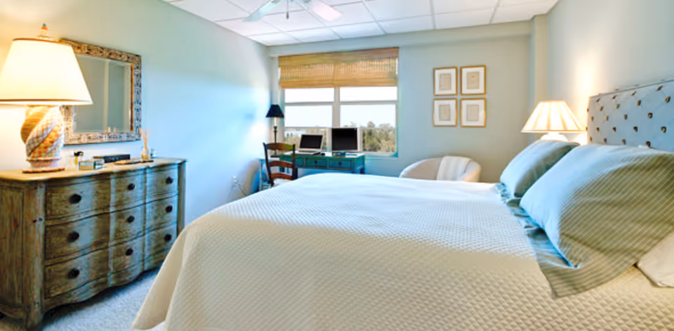 A bright bedroom with a large bed covered in a white quilt and two pillows. There is a wooden dresser with a decorative lamp and mirror on the left side. A window with a bamboo shade lets in natural light, and below it is a desk with two computer monitors and a chair. A small armchair and framed artwork are on the right wall.