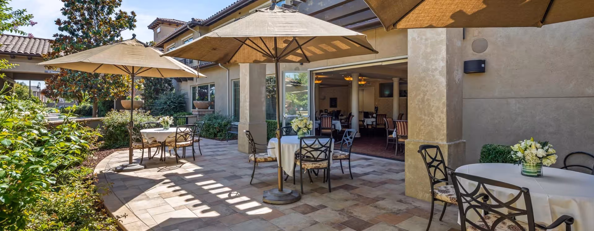 Outdoor patio area at Oakmont of San Antonio Heights with round tables covered in white tablecloths, each table surrounded by metal chairs with cushions. Large beige umbrellas provide shade over the tables. The patio is adjacent to a building with large windows and an open entrance leading to an indoor dining area. There are plants and greenery along the edge of the patio.