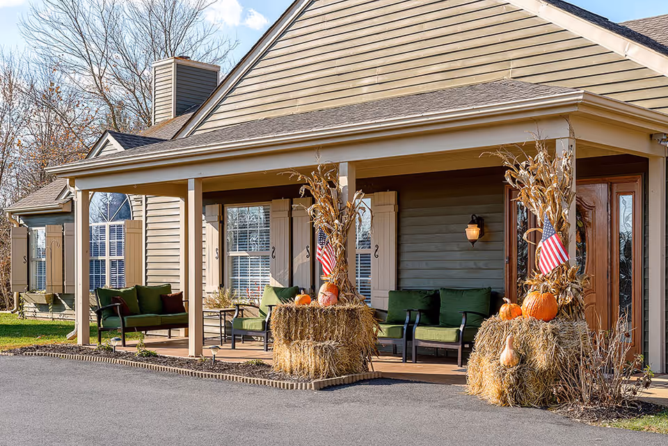 Front porch of a residential building decorated with hay bales, pumpkins, dried corn stalks, and small American flags. The porch has green cushioned chairs and a wooden door with a glass panel. The building exterior is beige with white trim and shuttered windows.