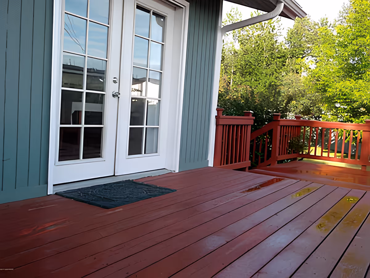 A wooden deck painted red with a black doormat in front of white double glass doors. The deck has a red railing and is surrounded by green trees and bushes.