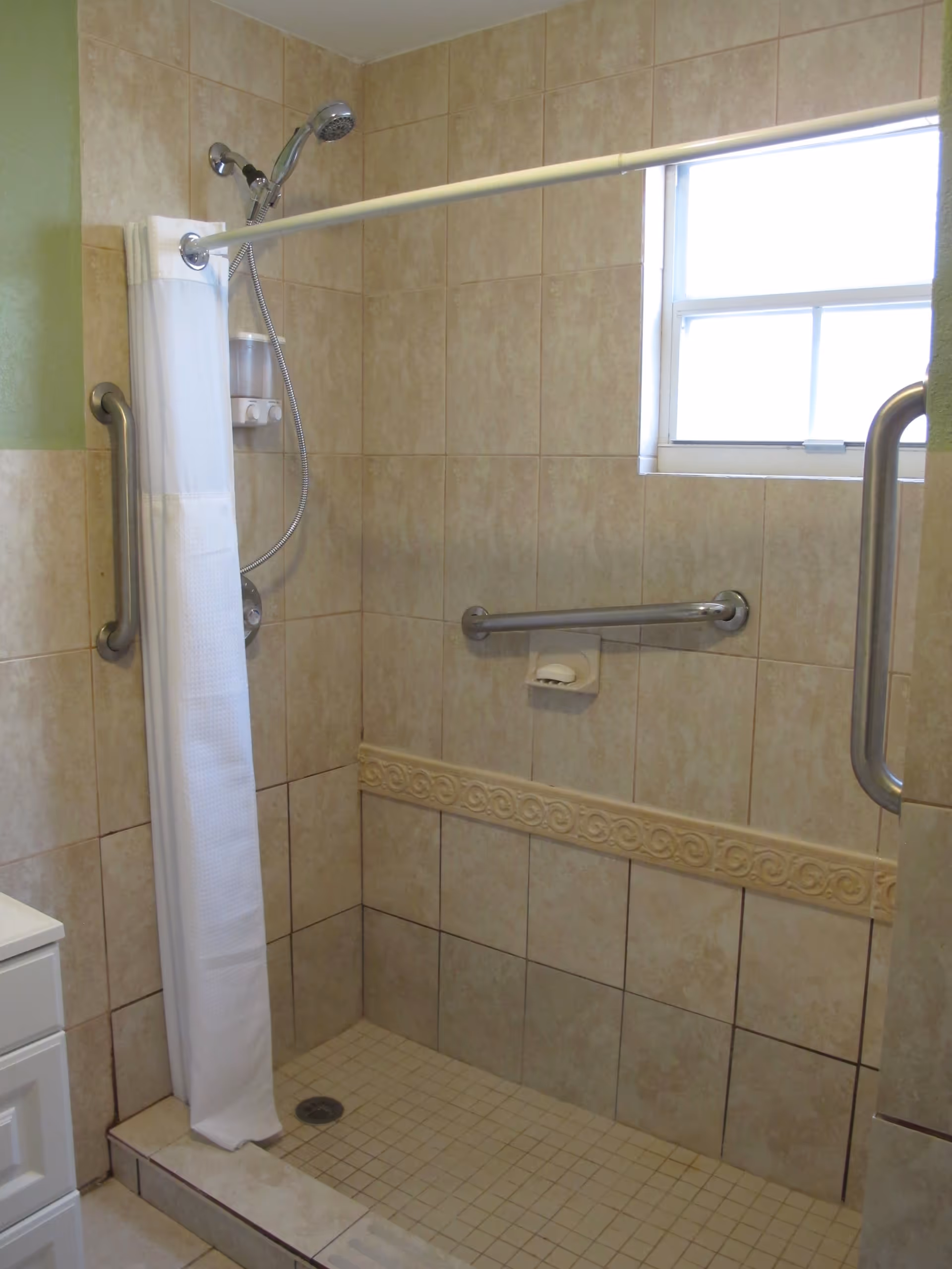 A tiled shower area with beige tiles and a decorative tile border. The shower has a handheld showerhead attached to a flexible hose, a white shower curtain hanging on a curved rod, and two stainless steel grab bars for support. There is a small window letting in natural light and a soap holder with a bar of soap.