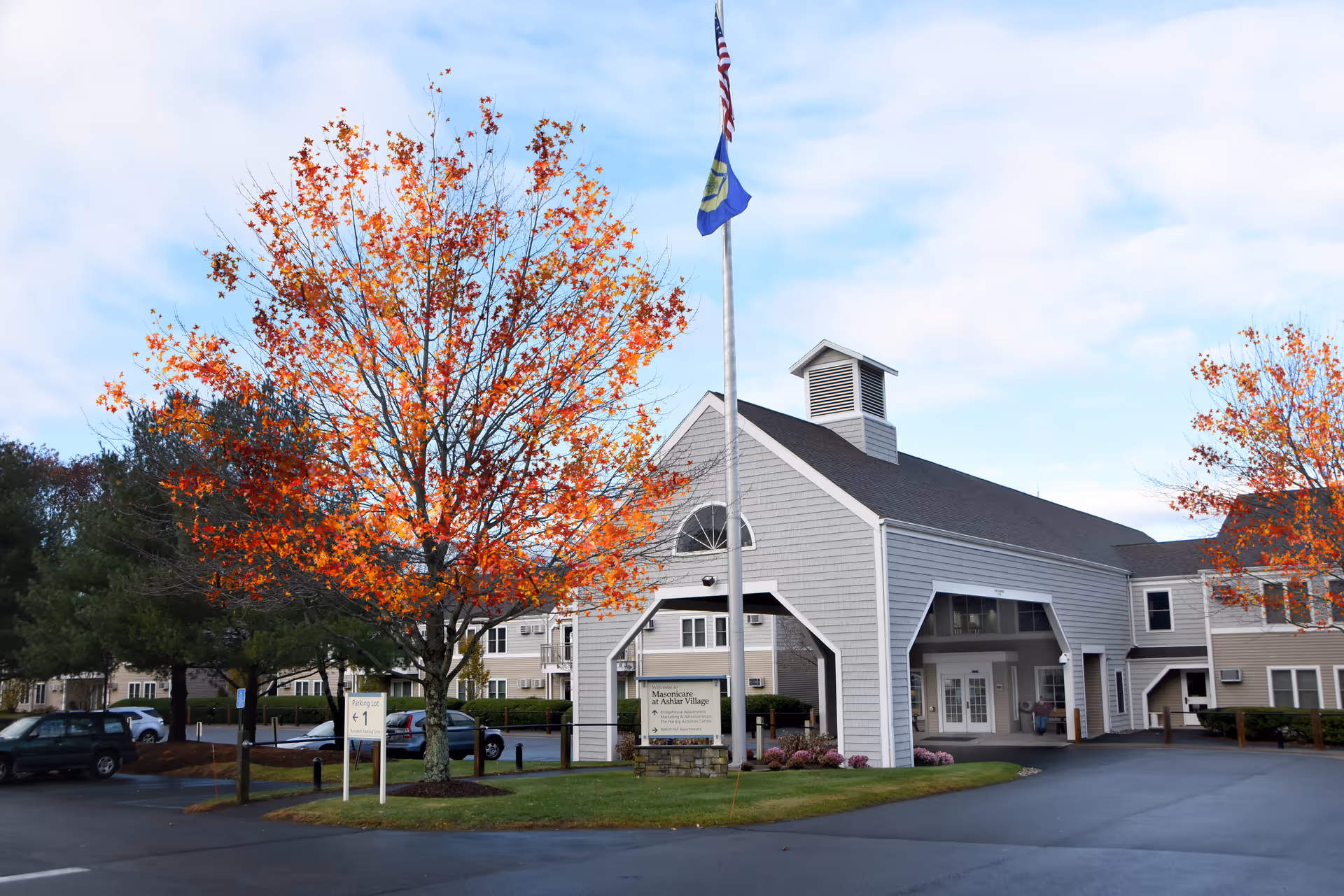 Exterior view of Masonicare At Ashlar Village facility with a gray building featuring a covered entrance, an American flag and another flag on a flagpole, and trees with autumn-colored leaves in the foreground.