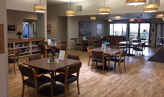 A spacious dining room with multiple wooden tables and chairs arranged neatly. Each table has a small flower arrangement and condiments. The room has wooden flooring, soft lighting from ceiling fixtures, and large windows letting in natural light. There is a bookshelf with books and a painting on the wall.
