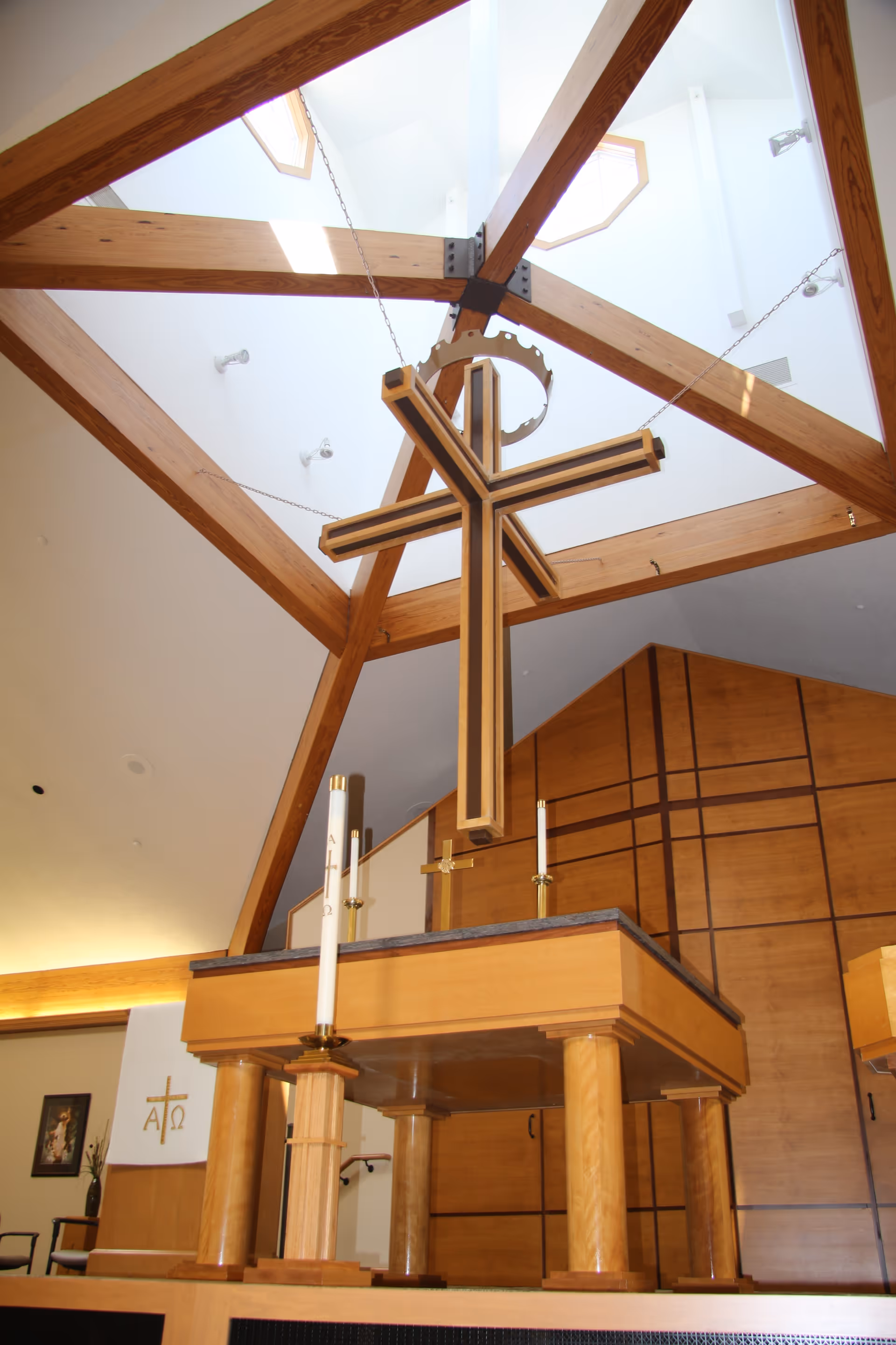 Interior view of a chapel with a large wooden cross suspended from the ceiling by chains. The ceiling has wooden beams and skylights. Below the cross is a wooden altar with candles. The walls and furniture are made of light-colored wood.