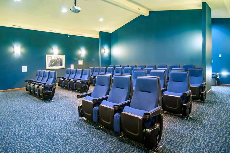 A small theater room with multiple rows of blue cushioned reclining seats arranged in front of a blank wall with a projector mounted on the ceiling. The walls are painted teal and have wall-mounted lights and a framed picture.