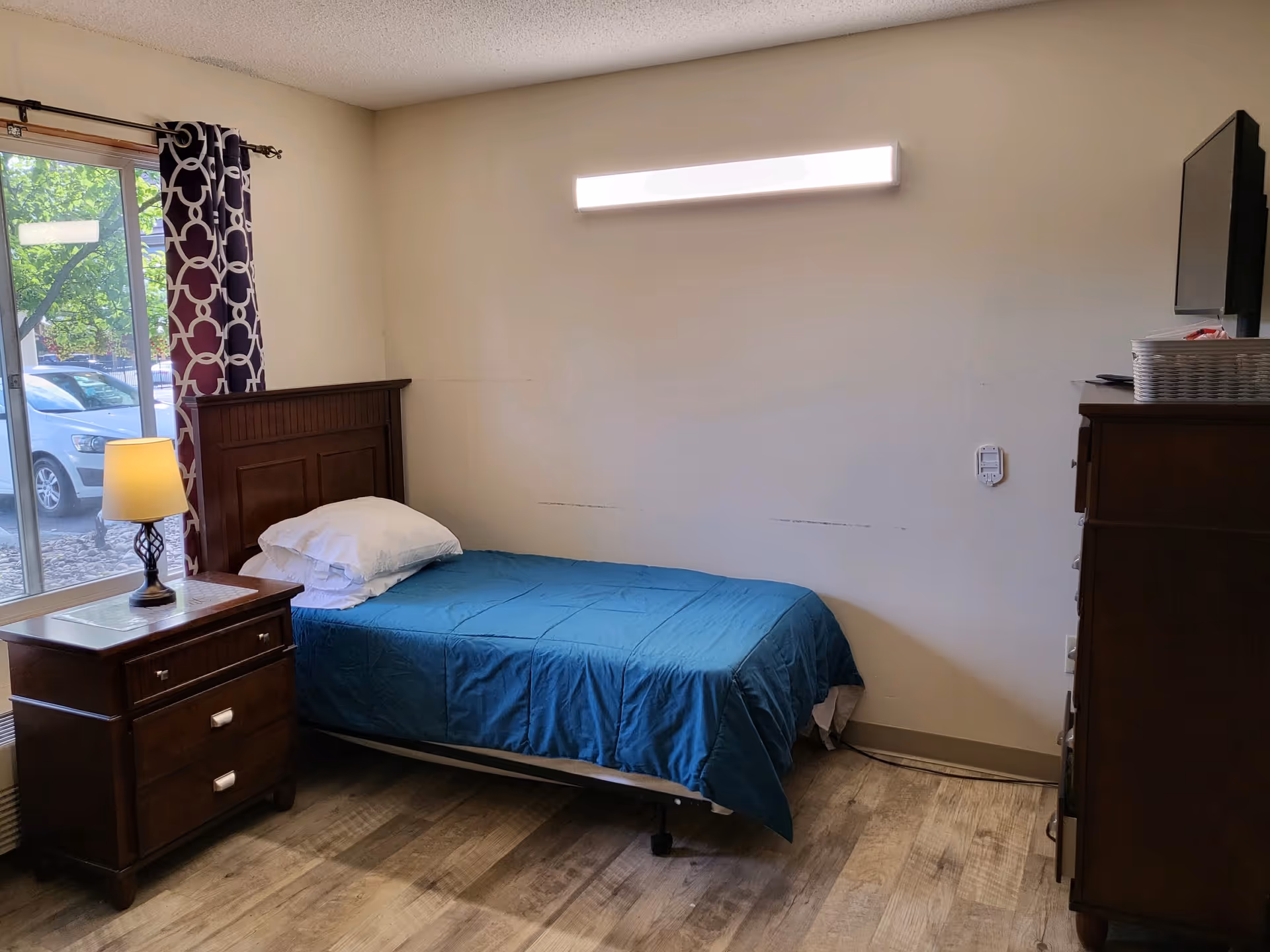 A simple bedroom with a single bed covered in a blue bedspread and white pillows. Next to the bed is a wooden nightstand with a lamp on it. A window with patterned curtains shows a view of a parked white car and greenery outside. On the right side, there is a wooden dresser with a TV on top. The room has light-colored walls and wood-patterned flooring.